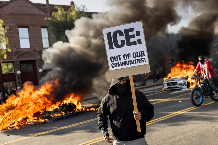 Protester holding sign against ICE amid fire and smoke during military crackdown in Los Angeles streets.