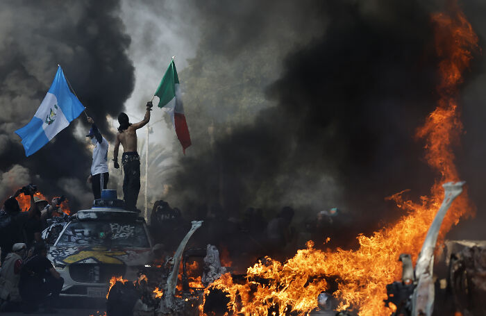 Protesters waving flags on burning cars amid thick smoke during LA riots linked to California Governor Newsom arrest talks.