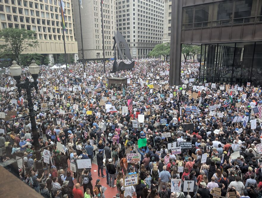 Large crowd of protesters gathered in urban area, holding signs during No Kings Rally with focus on fashion designer tribute.