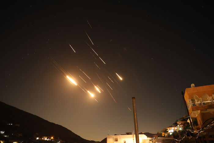Rockets blazing toward Israel at night while crowds calmly watch and celebrate on rooftops below the dark sky.