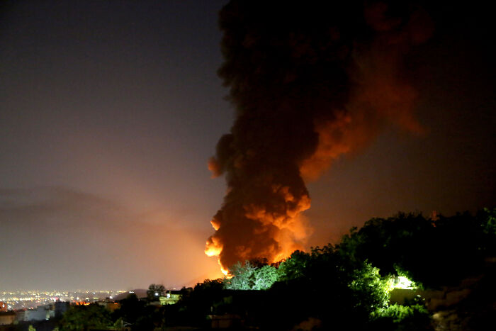 Smoke and fire blaze into the night sky as crowds stay calm on rooftops amid rockets toward Israel.