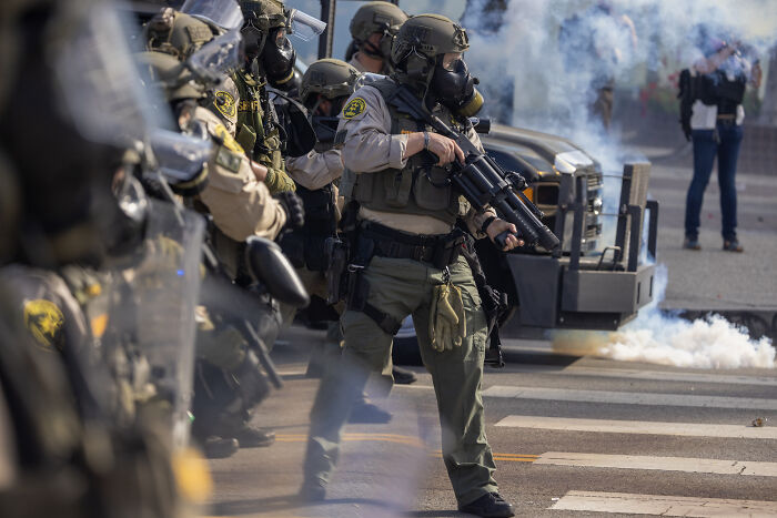 National Guard in tactical gear deployed in Los Angeles amid anti-ICE protests with smoke visible around them.