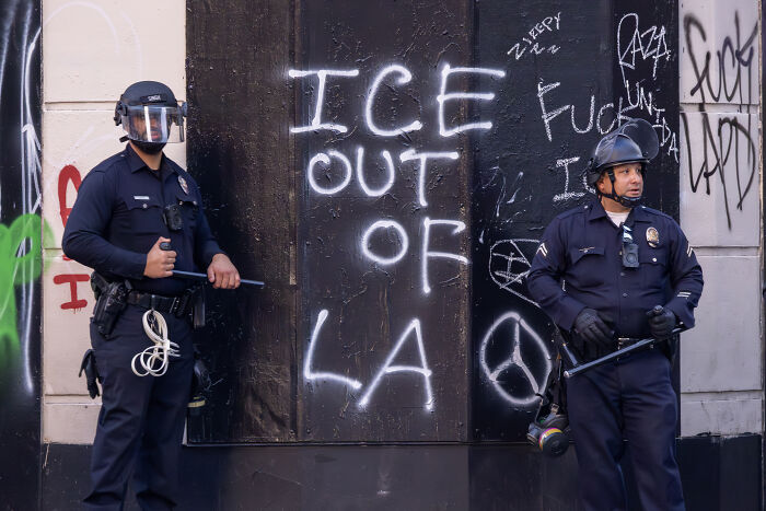 Two police officers in riot gear stand near graffiti reading ICE out of LA, related to California official and ICE raids.