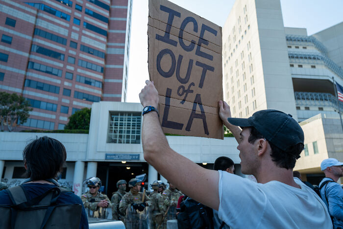 Man holding a sign saying ICE out of LA during a protest near police and military in a city setting in Los Angeles Dodgers area.