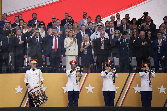 Military band performing at a parade with people standing and saluting during a political event featuring Trump and rally banners.