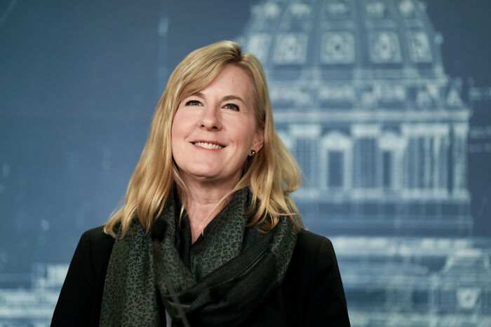Smiling woman with blonde hair and dark scarf standing in front of a blurred government building backdrop about alleged shooter in lawmakers attack.