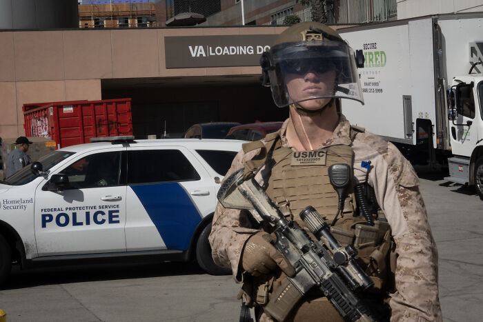 Armed USMC officer in tactical gear standing near Homeland Security police vehicle in Los Angeles Dodgers stadium parking lot.