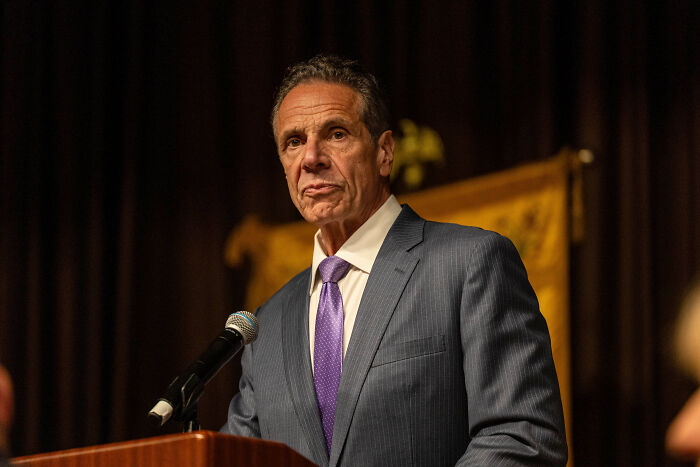 Man in suit and purple tie speaking at podium, representing Democratic Socialist Zohran Mamdani's NYC mayoral primary victory.