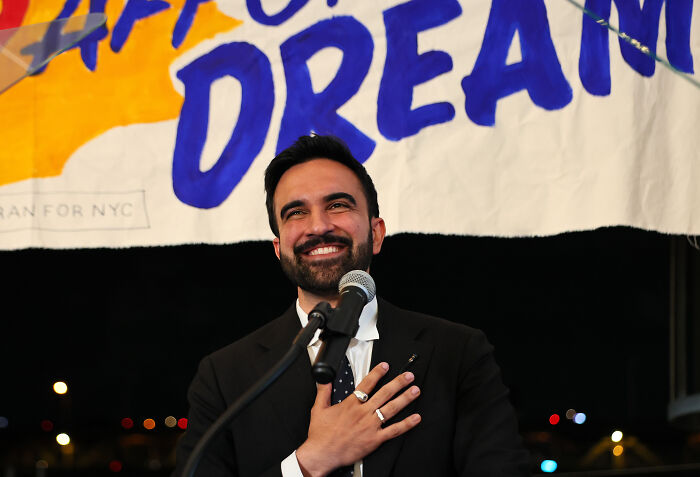 Democratic Socialist Zohran Mamdani smiling and speaking at a podium during NYC mayoral primary victory celebration.