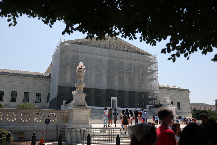 Exterior view of the Supreme Court building covered in scaffolding with visitors walking nearby on a clear day.