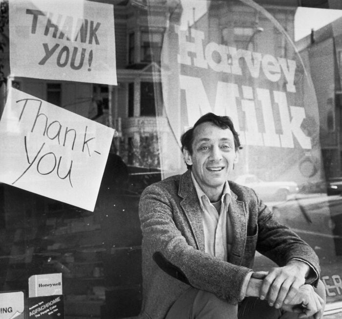 Black and white photo of Harvey Milk sitting outside a storefront with thank you signs, related to Navy rename vessel debate.