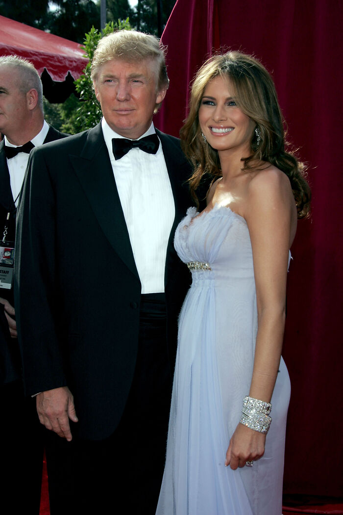 Donald Trump and Melania Trump dressed formally, posing together at an event with a red curtain backdrop.