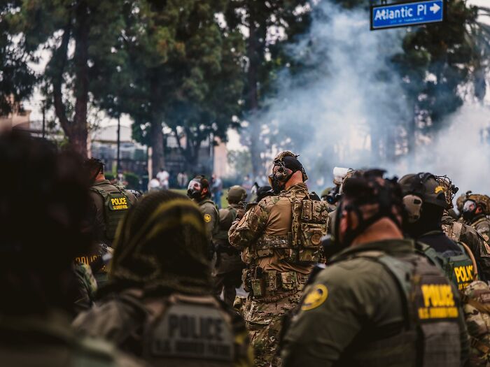 Heavily armed police officers in tactical gear during LA riots with smoke in the background and street sign visible.