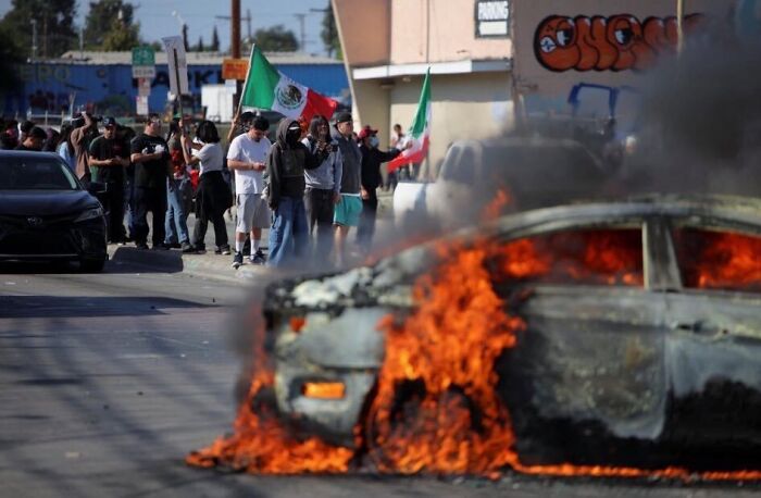 Car engulfed in flames during LA riots with protesters nearby amid calls for arrest of California Governor Gavin Newsom.