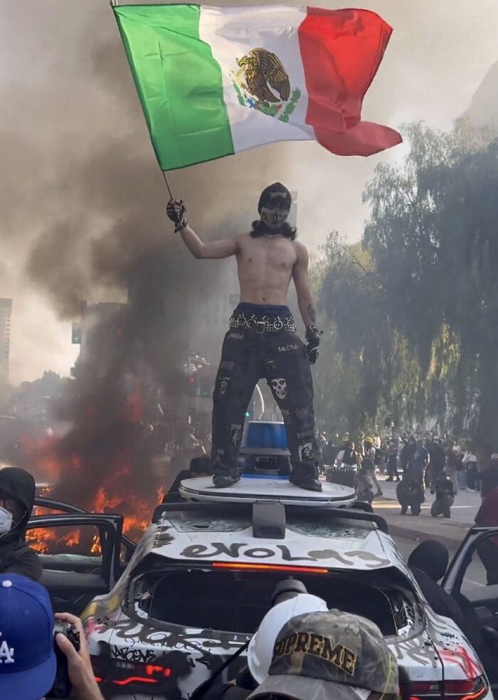 Protester stands on damaged police car holding flag amid chaos and smoke during LA riots with heavily armed cops present.
