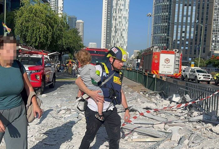 Rescue worker carries child amid rubble after Iran attack with emergency vehicles and city buildings in background.