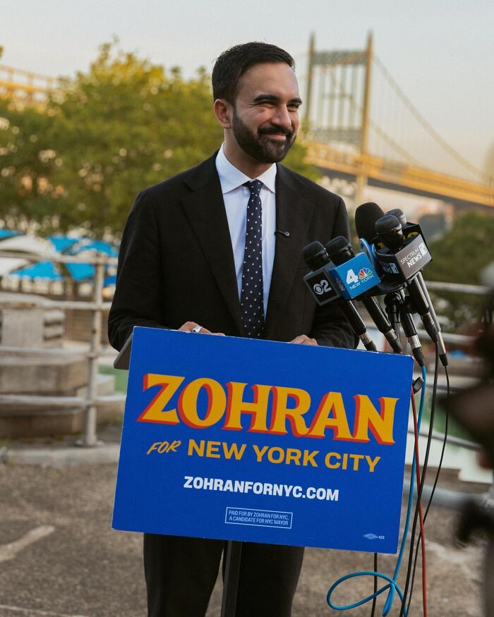 Democratic Socialist Zohran Mamdani smiling at a podium with microphones during NYC mayoral primary victory speech.