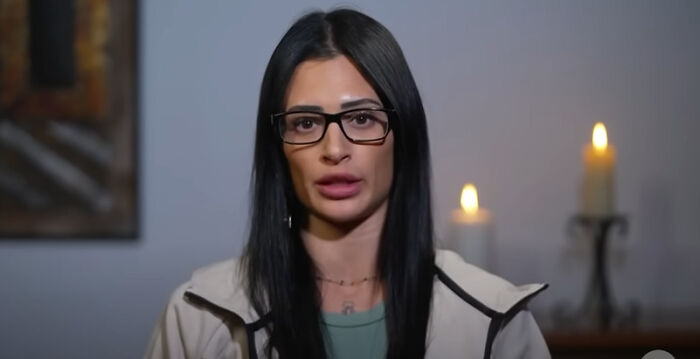 Young woman with black hair and glasses speaking indoors with lit candles in the background responding to DHS agents incident.