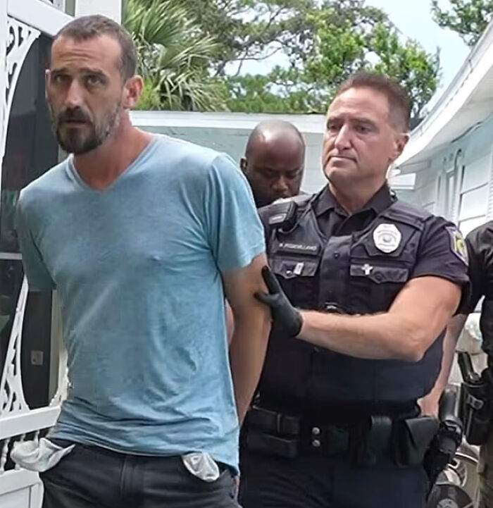 Man in blue shirt being escorted by police officers outside a home in Florida linked to baby boy dies after father leaves him in boiling car.