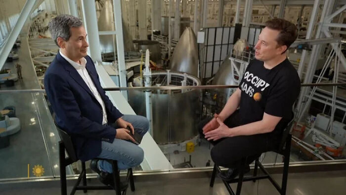 Elon Musk seated during an interview, discussing topics related to foreign student ban and spaceships in a modern facility.