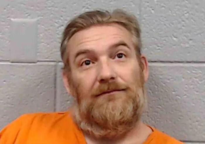 Man charged with sexually abusing daughter for 12 years, wearing orange prison uniform, against gray block wall background.