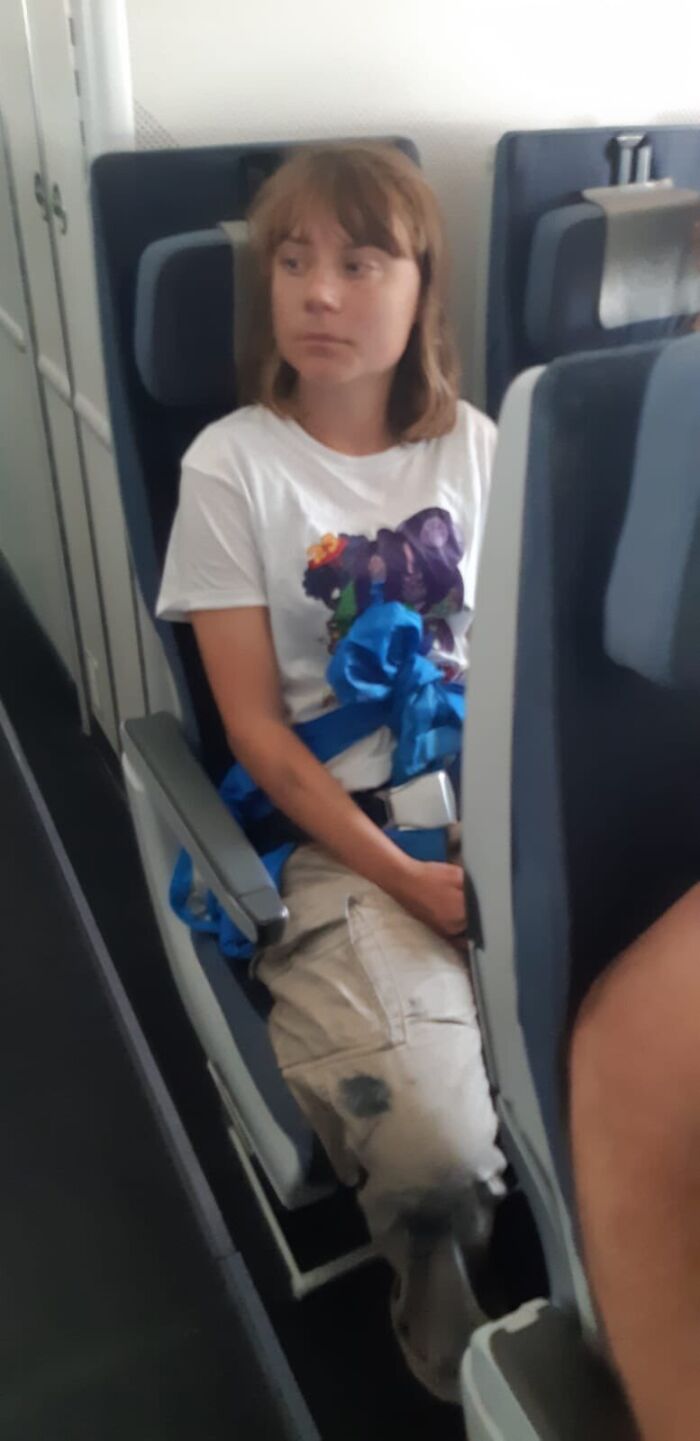 Greta Thunberg seated on a plane, wearing a white graphic t-shirt and cargo pants during Israel deportation after Gaza aid blockade attempt
