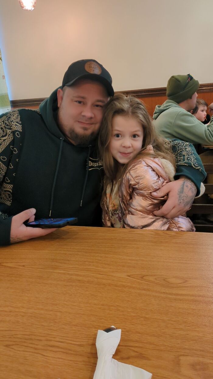 Man in black hoodie and cap embracing young girl in pink jacket sitting at a wooden table indoors with other people.
