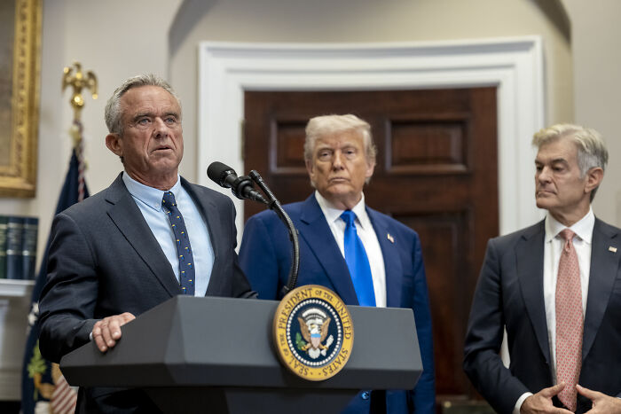Man speaking at presidential podium with Donald Trump and two men listening in a formal government setting about neocon war hawk.
