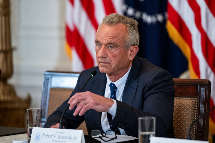 Robert F. Kennedy Jr. speaking at a formal event with American flags in the background, discussing politics.