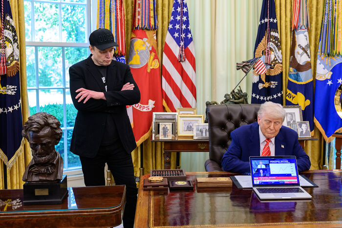 Elon Musk standing with arms crossed while former President Trump sits at Resolute Desk with laptop in the Oval Office.