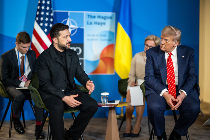 Ukrainian President and Donald Trump seated during a meeting with NATO and Ukraine flags in the background.