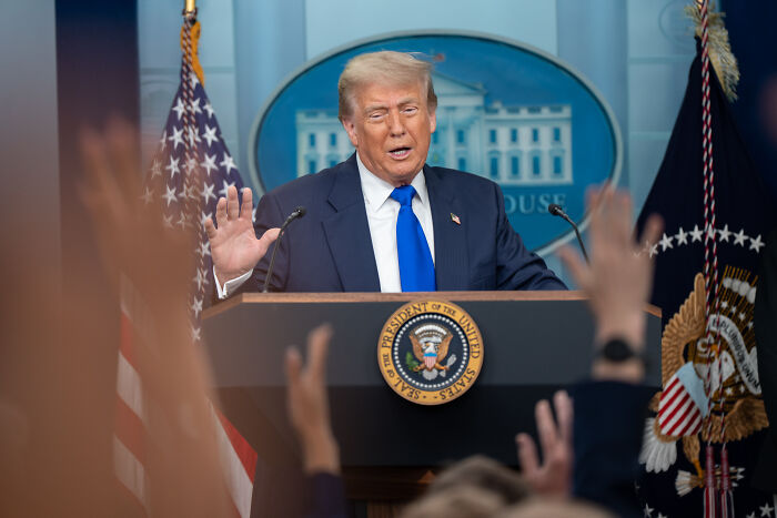 Donald Trump speaking at a podium in the White House press room as reporters raise hands during Q&A session.