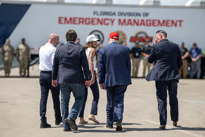 Group of people, including Trump wearing a red cap, walking near Florida Division of Emergency Management building.