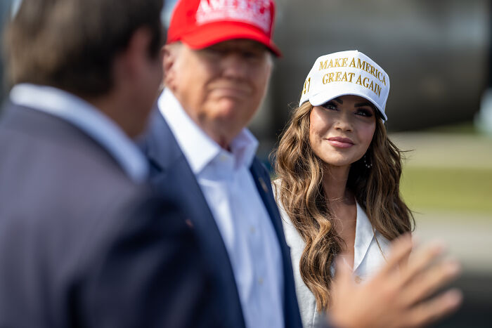 Donald Trump and Kristi Noem wearing MAGA hats, engaging in a conversation outdoors at a political event.