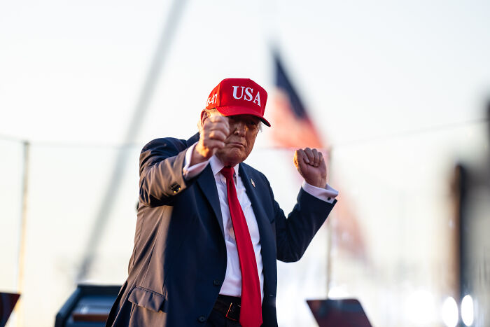 Former President Trump wearing a red USA hat, gesturing emphatically during a public event with an American flag in the background.
