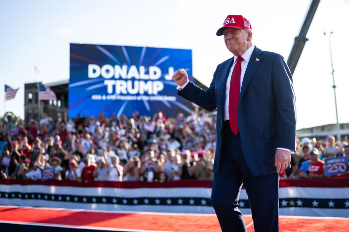 Former President Donald Trump at a rally, wearing a red USA hat and blue suit, with a crowd and stage in the background.