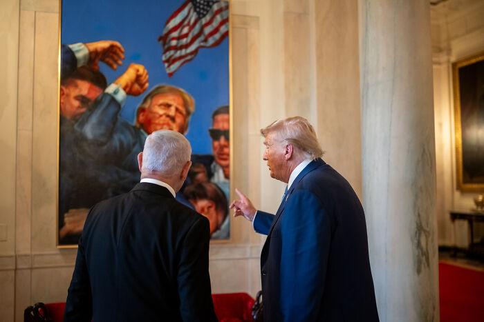 Netanyahu and Trump stand indoors near a large painting with an American flag, symbolizing peace prize nomination.