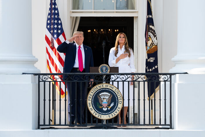 Donald Trump and Melania Trump on White House balcony with presidential seal and US flags during salute and pledge.
