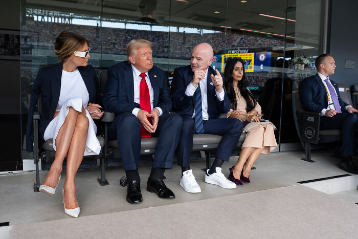 Donald Trump attending event wearing suit, seated between two people, showing signs of swollen legs in public setting.