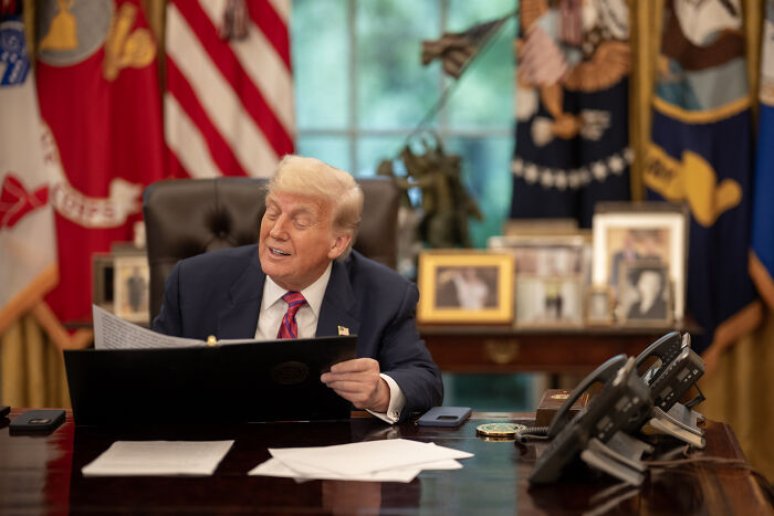 Donald Trump sitting at the Resolute Desk in the Oval Office, holding a document, with flags in the background.
