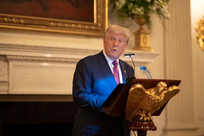 Donald Trump speaking at a podium with a golden eagle emblem, relating to anti-trans agenda coverage.