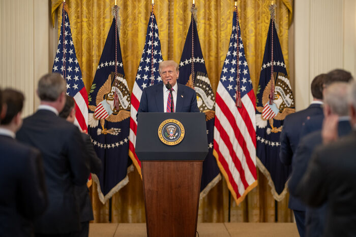 Former President Trump speaking at a podium with presidential seal during a formal event with American flags in the background.