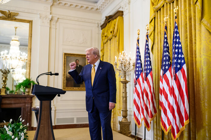 Donald Trump speaking at a podium in a formal room with multiple American flags and gold curtains behind him.