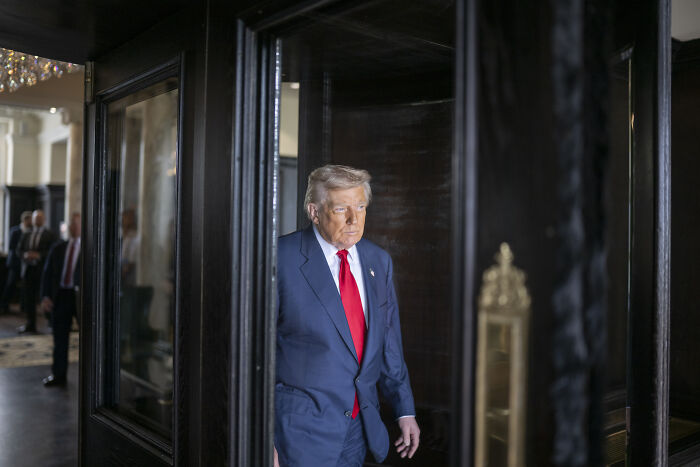 Donald Trump in a navy suit and red tie walking indoors, with a serious expression amid a formal setting.