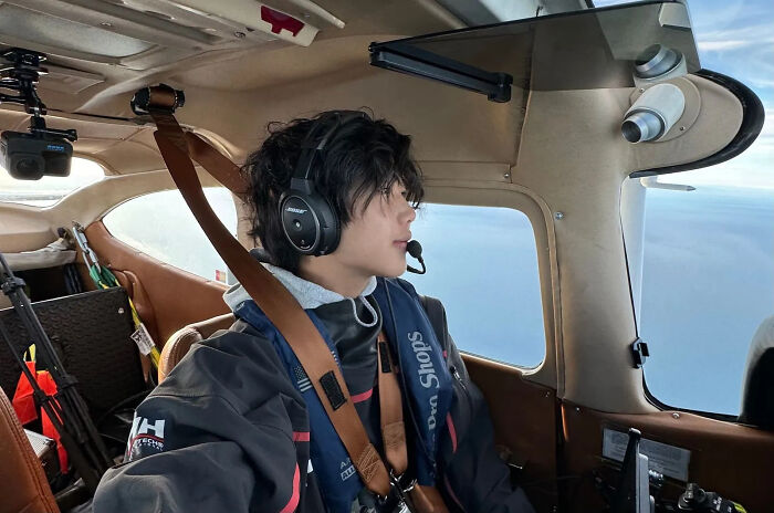US teen pilot wearing headset and jacket inside small aircraft cabin during solo flight supporting cancer cause.