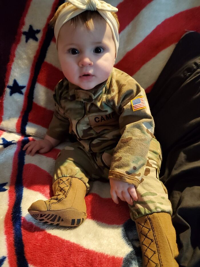 One-year-old baby girl dressed in camouflage outfit sitting on a blanket with stars and stripes pattern.