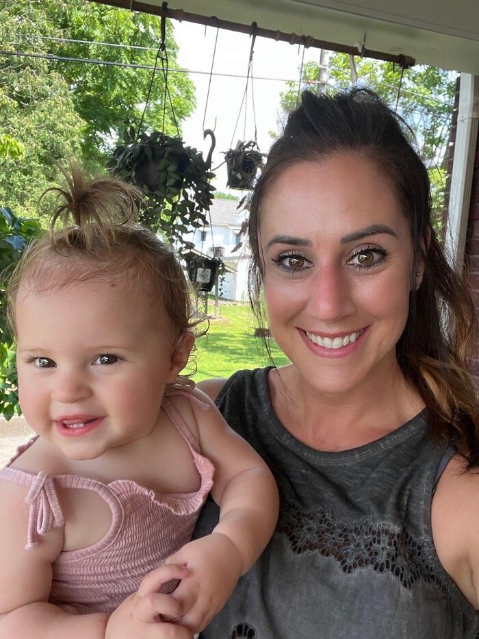 Ohio mother and one-year-old baby girl smiling outdoors on a porch with hanging plants in the background.