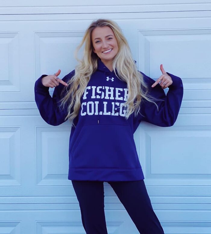 Young woman wearing a Fisher College sweatshirt, smiling and pointing at the logo in front of a white garage door