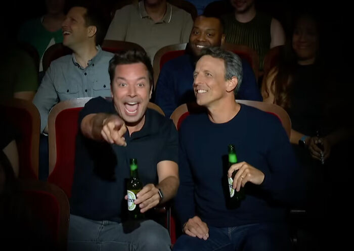 Audience members laughing and holding drinks in a theater, highlighting Trump's quest to sue and silence TV critics.