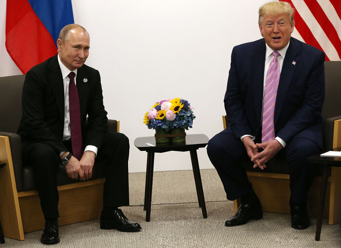 Vladimir Putin and Donald Trump seated in a formal meeting room with flowers on a small table between them.